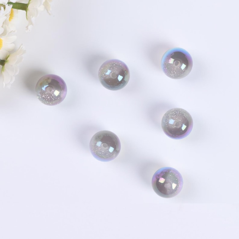 May include: Six iridescent glass beads with a glittery finish. The beads are round and have a hole in the center. They are a light purple color with a hint of blue.