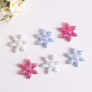 Peut inclure: Six ornements en forme de flocons de neige irisés, dans les tons de blanc, rose et bleu. Les pièces décoratives sont disposées sur une surface blanche, avec quelques fleurs blanches en arrière-plan. Idéal pour les projets d'artisanat.