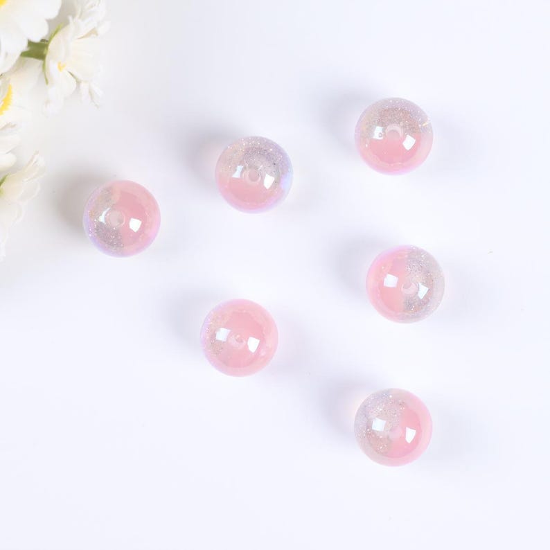 May include: Six iridescent pink glass beads with a hole in the center. The beads are scattered on a white surface.