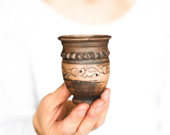 May include: A small, handcrafted ceramic vase with a dark brown exterior and a lighter tan band featuring an etched floral design. The vase has a decorative rim and a rounded base, held against a white background.