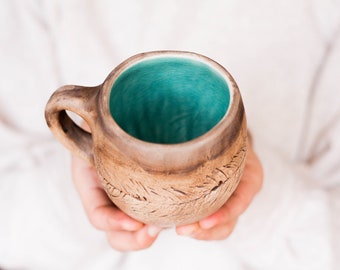 May include: A brown ceramic mug with a teal blue glaze inside. The mug has a textured, hand-crafted look with a design of lines and swirls.