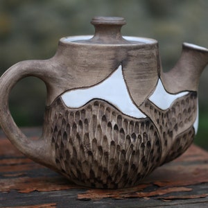 Può includere: Una teiera in ceramica marrone con un design bianco. La teiera ha una superficie testurizzata e un coperchio.