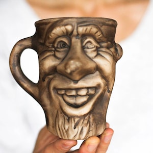 May include: A brown ceramic mug shaped like a smiling face with a large nose and a wide grin. The mug has a handle and is made to look like a cartoon character.