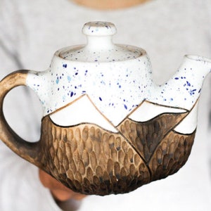 May include: White ceramic teapot with a brown mountain design and blue speckles. The teapot has a lid and a handle.