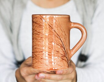 May include: A brown ceramic mug with a design of brown lines resembling feathers. The mug has a rounded handle.