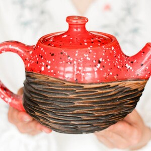 Può includere: Una teiera in ceramica rossa e marrone con macchie bianche. La teiera ha un fondo marrone strutturato e una parte superiore rossa liscia.