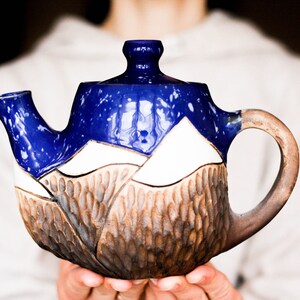 Può includere: Una teiera in ceramica blu e bianca con una base marrone testurizzata. La teiera ha un design a tema montagna e un manico arrotondato.