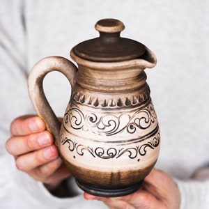 May include: A brown ceramic pitcher with a lid, featuring a white background and intricate brown floral designs. The pitcher has a handle and a spout.