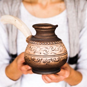 May include: A brown ceramic coffee pot with a decorative design and a white handle. The pot is being held by a person's hands.