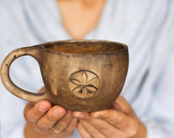 May include: A brown ceramic mug with a carved floral design. The mug is being held by a person's hands.