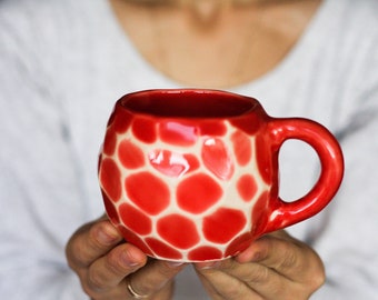 May include: A red ceramic mug with a white honeycomb pattern. The mug has a rounded body and a handle.