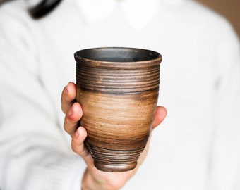 May include: A handmade ceramic cup with a dark brown interior and a lighter brown exterior. The cup has horizontal ridges and is held in a person's hand. The cup is approximately 4 inches tall.