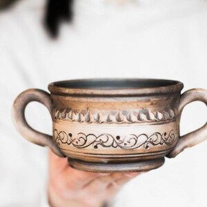 Peut inclure: Une tasse en céramique brune avec un motif décoratif de volutes et de lignes. La tasse a deux anses et une forme arrondie.