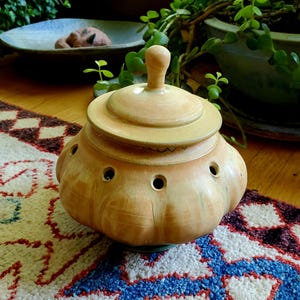 May include: A round, light tan ceramic garlic keeper with a matching lid and a knob handle. The vessel features a series of small, evenly spaced circular ventilation holes around the upper shoulder area and a smooth, glazed finish throughout.
