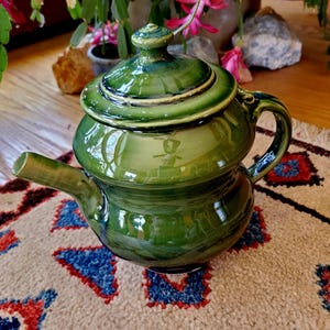 May include: A glossy, green ceramic teapot with a lid and spout. The teapot has a rounded body and a handle. The background includes a patterned rug and plants with pink flowers. The teapot is a decorative item.