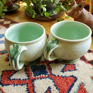 May include: Two ceramic mugs with a pale mint green interior and a white glazed exterior. Each mug features a sculpted green frog perched on the handle. The mugs have a rounded, bowl-like shape with a smooth finish and a slightly flared rim.