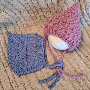 May include: Two knitted baby bonnets, one blue and one pink, with ties. The bonnets are lying on a beige knitted blanket.