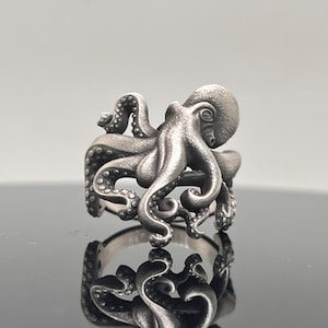 Peut inclure: Une bague argentée en forme de poulpe. La bague présente un motif de poulpe détaillé avec des tentacules enroulées autour de l'anneau et une tête de poulpe centrale. La bague est présentée sur une surface réfléchissante.