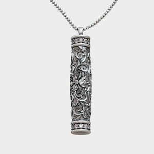Puede incluir: Collar con colgante de plata con un colgante cilíndrico con intrincados diseños florales y de volutas. El colgante está adornado con pequeñas piedras preciosas transparentes y cuelga de una delicada cadena de cuentas.
