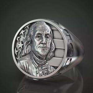 Könnte beinhalten: Ein silberner Ring mit einem detaillierten Relief von Benjamin Franklin. Die Ringoberfläche zeigt Sterne und Streifen, mit dem Profil von Franklin in der Mitte. Der Ring hat eine polierte Oberfläche und ein abgerundetes Band.