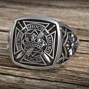 May include: A silver-colored ring with a square face featuring a cross, crown, and swords design. The ring has the words "IN HOC SIGNO VINCES" and "HOC" engraved on it. The ring is set on a wooden surface.