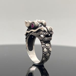Puede incluir: Anillo de dragón plateado con detalles intrincados. La cabeza del dragón presenta un ojo de gema roja. El diseño del anillo incluye escamas y una cara detallada, mostrando un tema mítico.