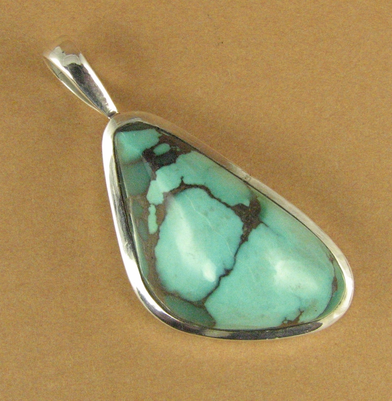 May include: A turquoise pendant with a silver setting. The turquoise is a light blue-green color with brown veins.