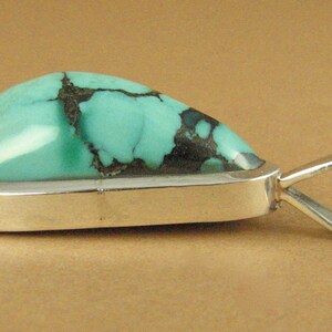 May include: A turquoise pendant with a silver setting and a bail. The turquoise is a light blue-green color with black veins.