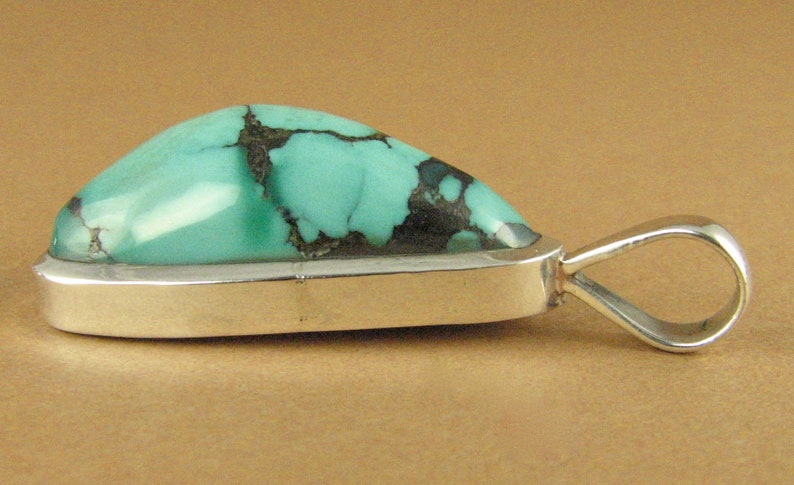 May include: A turquoise pendant with a silver setting and a bail. The turquoise is a light blue-green color with black veins.
