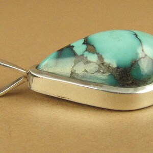 May include: A silver pendant with a teardrop-shaped turquoise stone. The stone has a green and blue color with black and white veins.