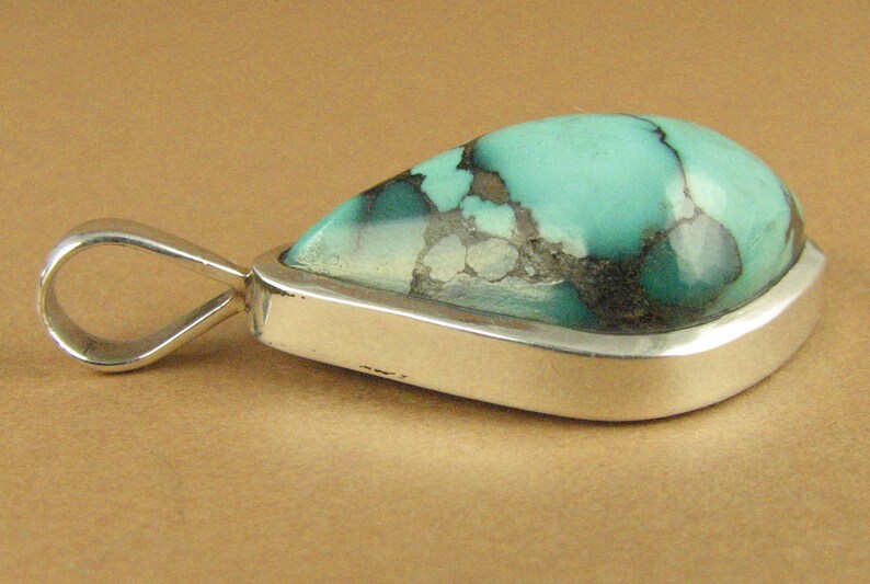 May include: A silver pendant with a teardrop-shaped turquoise stone. The stone has a green and blue color with black and white veins.