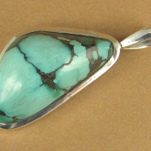 May include: A turquoise pendant with a silver setting. The turquoise is a light blue-green color with dark brown veins.