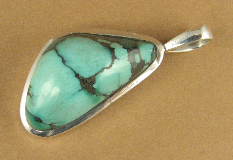 May include: A turquoise pendant with a silver setting. The turquoise is a light blue-green color with dark brown veins.