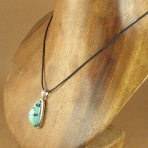 May include: A turquoise pendant necklace with a silver setting on a black cord. The necklace is displayed on a wooden mannequin.