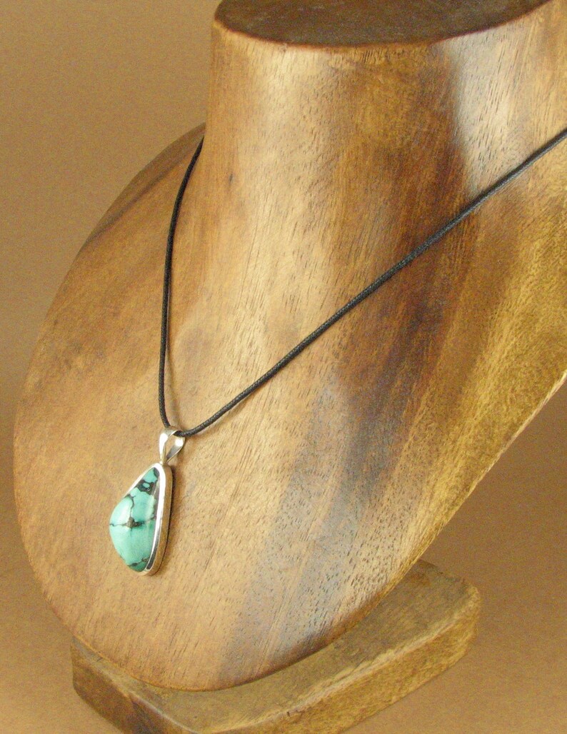May include: A turquoise pendant necklace with a silver setting on a black cord. The necklace is displayed on a wooden mannequin.