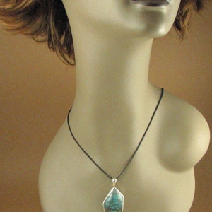 May include: A turquoise gemstone pendant with a silver setting hangs from a black cord necklace. The pendant is a geometric shape with a dark green and blue color.