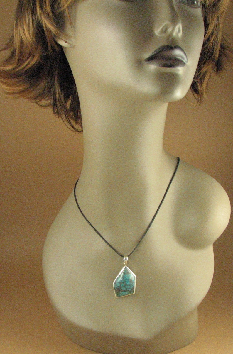 May include: A turquoise gemstone pendant with a silver setting hangs from a black cord necklace. The pendant is a geometric shape with a dark green and blue color.