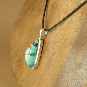 May include: A silver pendant with a turquoise stone set in a teardrop shape. The pendant is hanging from a black cord.