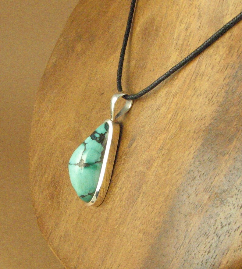 May include: A silver pendant with a turquoise stone set in a teardrop shape. The pendant is hanging from a black cord.