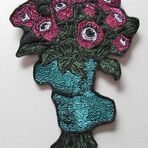 May include: Embroidered patch depicting a bouquet of pink roses with white detailing and green foliage, set against a black backdrop. The flowers are held in a teal vase-shaped design, offering a vivid contrast.