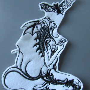 May include: Black and white embroidered patch featuring a woman with wings and a tail, kneeling with her head tilted back. A bat flies above her.