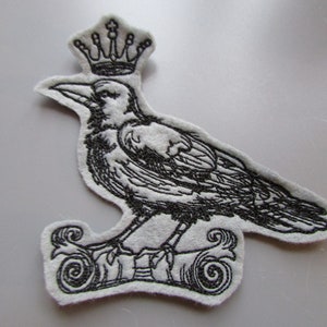 Könnte beinhalten: Schwarz-weiß gestickter Bügelaufkleber mit einer Krönchen tragenden Krähe, die auf einer Schriftrolle sitzt.