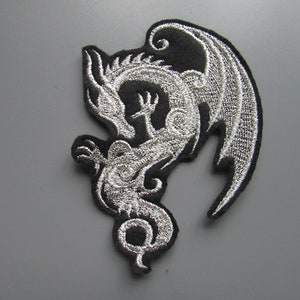 May include: A silver embroidered patch of a dragon with wings outstretched, on a black background.