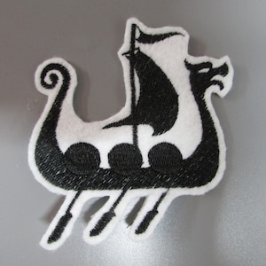 May include: Embroidered patch depicting a black and white Viking ship. The ship has a dragon head, a sail, and three round portholes. The patch is designed for application on clothing or accessories.