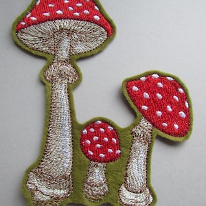 May include: Embroidered patch featuring three red and white mushrooms with white spots on a green felt background.