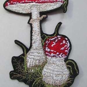 May include: Embroidered patch featuring two red and white mushrooms with white stems and green foliage. The patch is outlined in black.