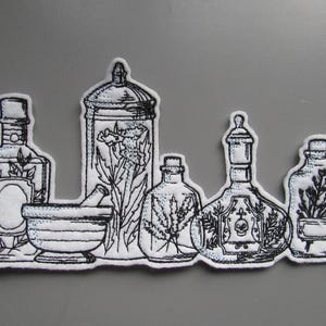 May include: A set of six black and white embroidered apothecary bottles and a mortar and pestle. The bottles are all different shapes and sizes and have various herbs and flowers inside. The bottles are on a white background.