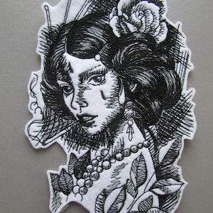 May include: Black and white embroidered patch of a woman with a rose in her hair, wearing a necklace and earrings. The woman's face is partially obscured by her hair. The background is a stylized design of leaves and branches.