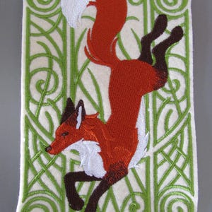 May include: Embroidered patch depicting a leaping fox with a white-tipped tail. The fox is mainly orange with white and brown details. The patch has a cream background with green decorative elements.
