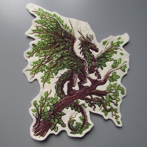 May include: An embroidered patch featuring a dark purple dragon perched on a brown tree branch with vibrant green leaves. The design is stitched onto a cream-coloured fabric backing with an irregular, die-cut edge that follows the shape of the art.